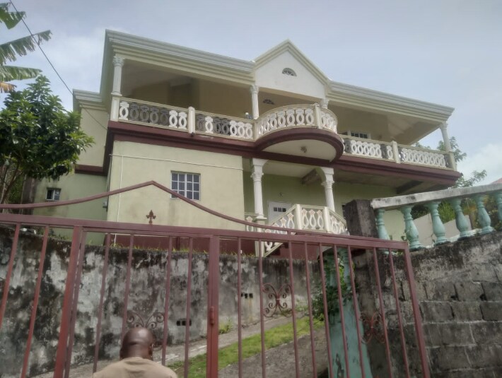 A large, two-story building is surrounded by greenery, featuring a decorative balcony and a front gate. The exterior walls are painted in a light color, while the structure is characterized by architectural details around the windows and doors.