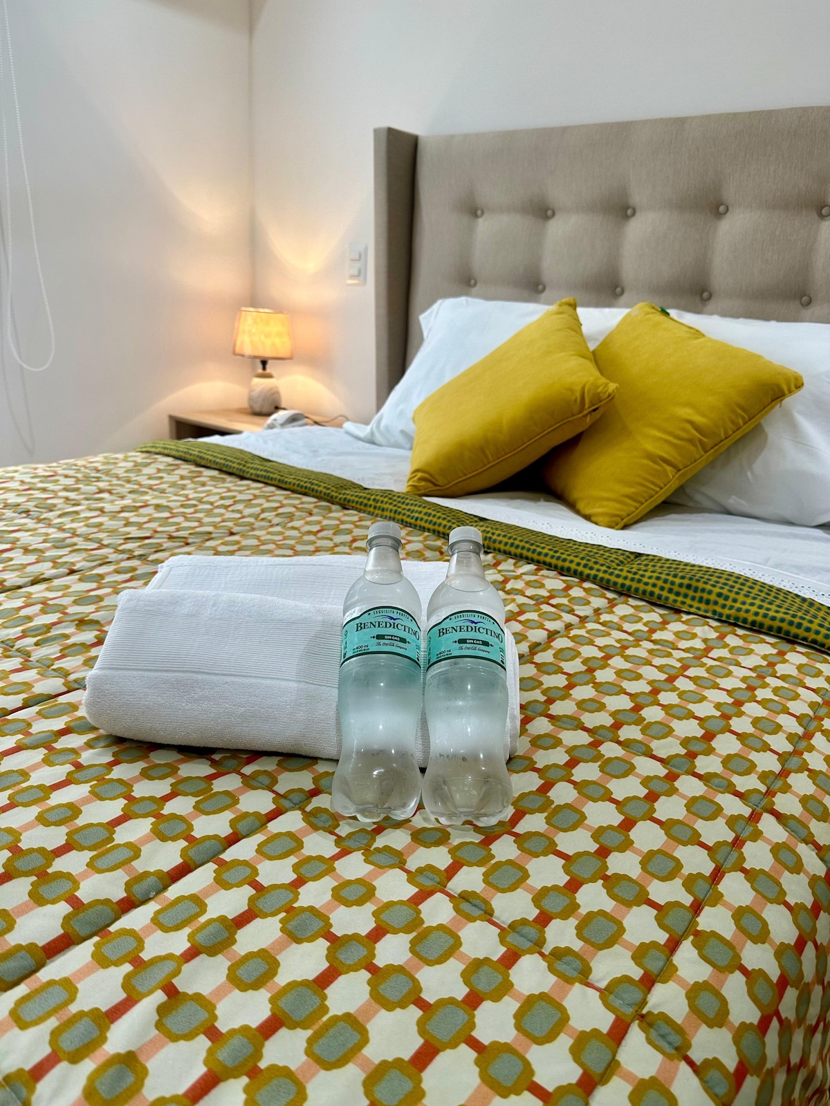 A neatly arranged sleeping area features a spacious bed adorned with yellow accent pillows against a patterned bedspread. Two bottles of water are placed beside a neatly folded white towel, while a lamp on the bedside table adds a soft glow to the room.