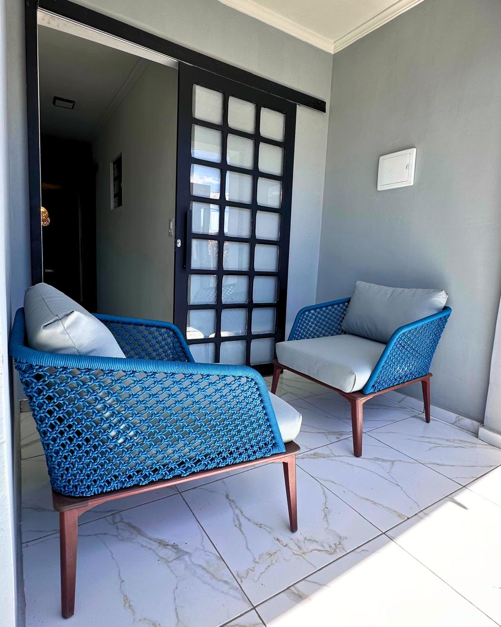A small seating area features two modern blue woven chairs with light cushions placed on a tiled floor. A glass-paneled door leads to a hallway, allowing natural light to fill the space.