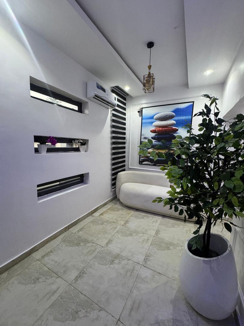 A bright hallway features a serene landscape painting with stacked stones, positioned against a white wall. A decorative plant in a white pot adds a touch of greenery, while natural light highlights the light-colored tiles and modern air conditioning unit.