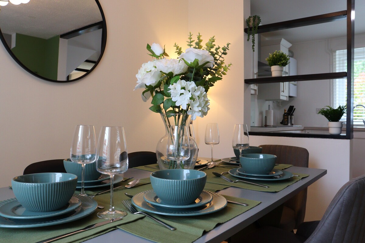 A dining area is set with green tableware, glassware, and chic cutlery, arranged thoughtfully on a textured tablecloth. A floral centerpiece adds a fresh touch, complemented by soft lighting and a neutral backdrop. The kitchen can be seen in the background, enhancing the inviting atmosphere.