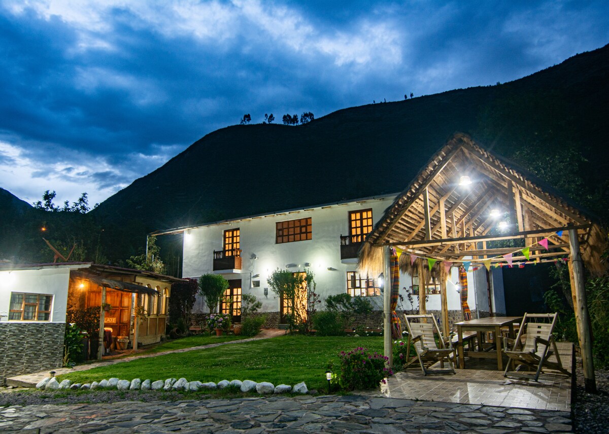 The serene lodge is set against a mountainous backdrop, featuring a rustic structure with multiple windows. A charming outdoor seating area under a thatched roof is illuminated, surrounded by lush greenery and garden spaces, inviting relaxation and social gatherings.