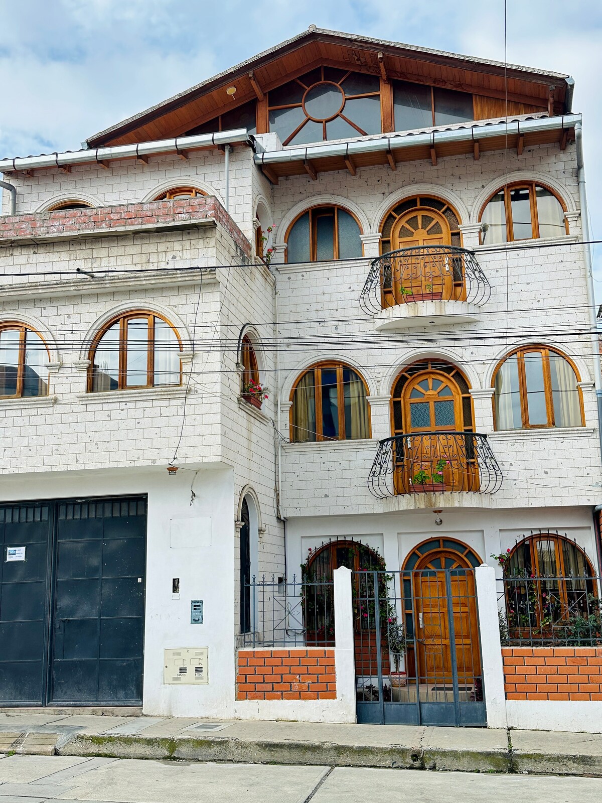 A charming multi-story building is presented, featuring distinctive round windows and a combination of white stone and brick exteriors. Balconies adorned with decorative railings are visible on each level, enhancing the building's architectural appeal. The entrance is framed by plants and flowers.