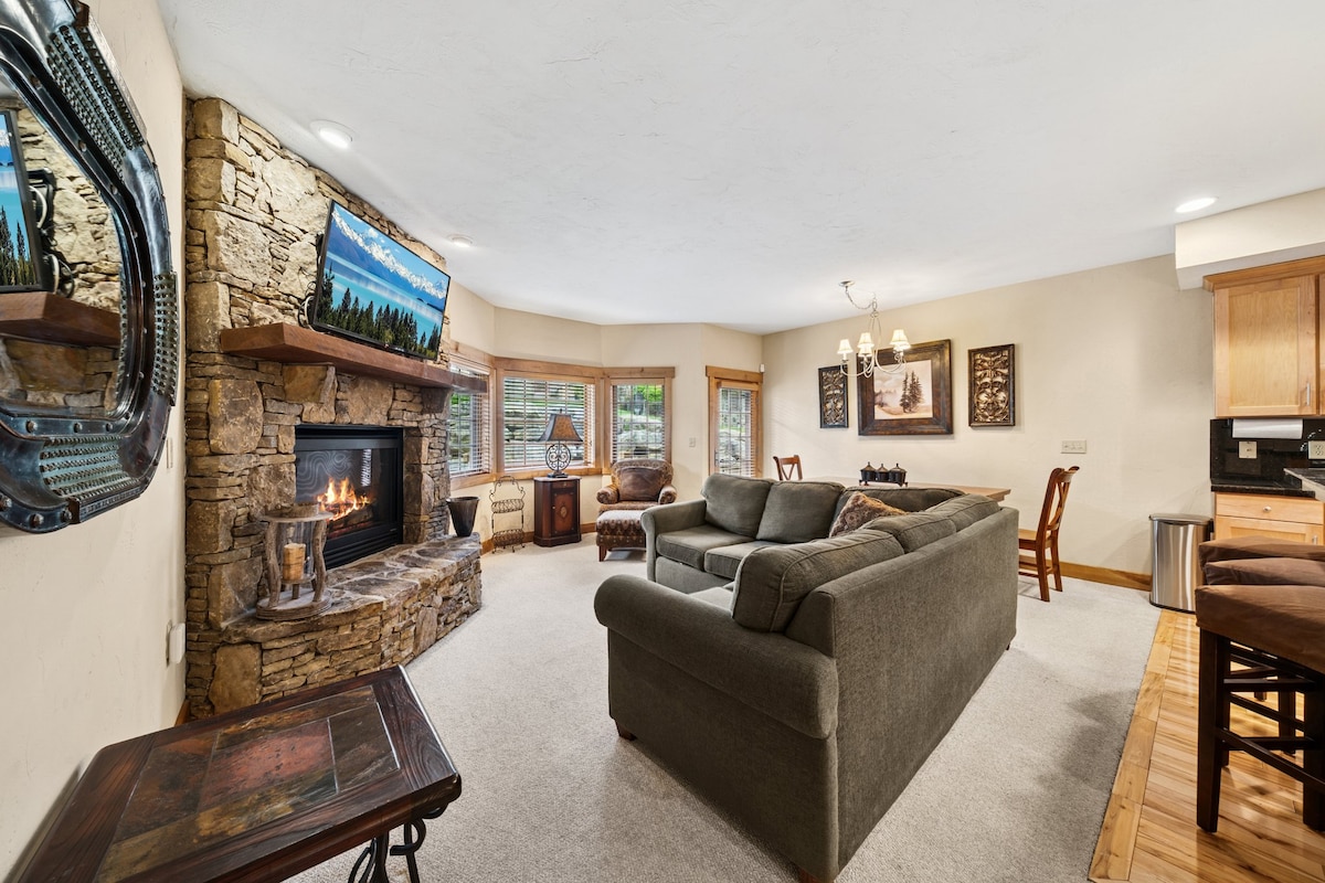 The cozy living room features a large sectional sofa facing a stone fireplace, with a wall-mounted TV above it. Windows allow natural light, and a dining table is set nearby. A rustic wooden coffee table sits in the center, enhancing the inviting space.