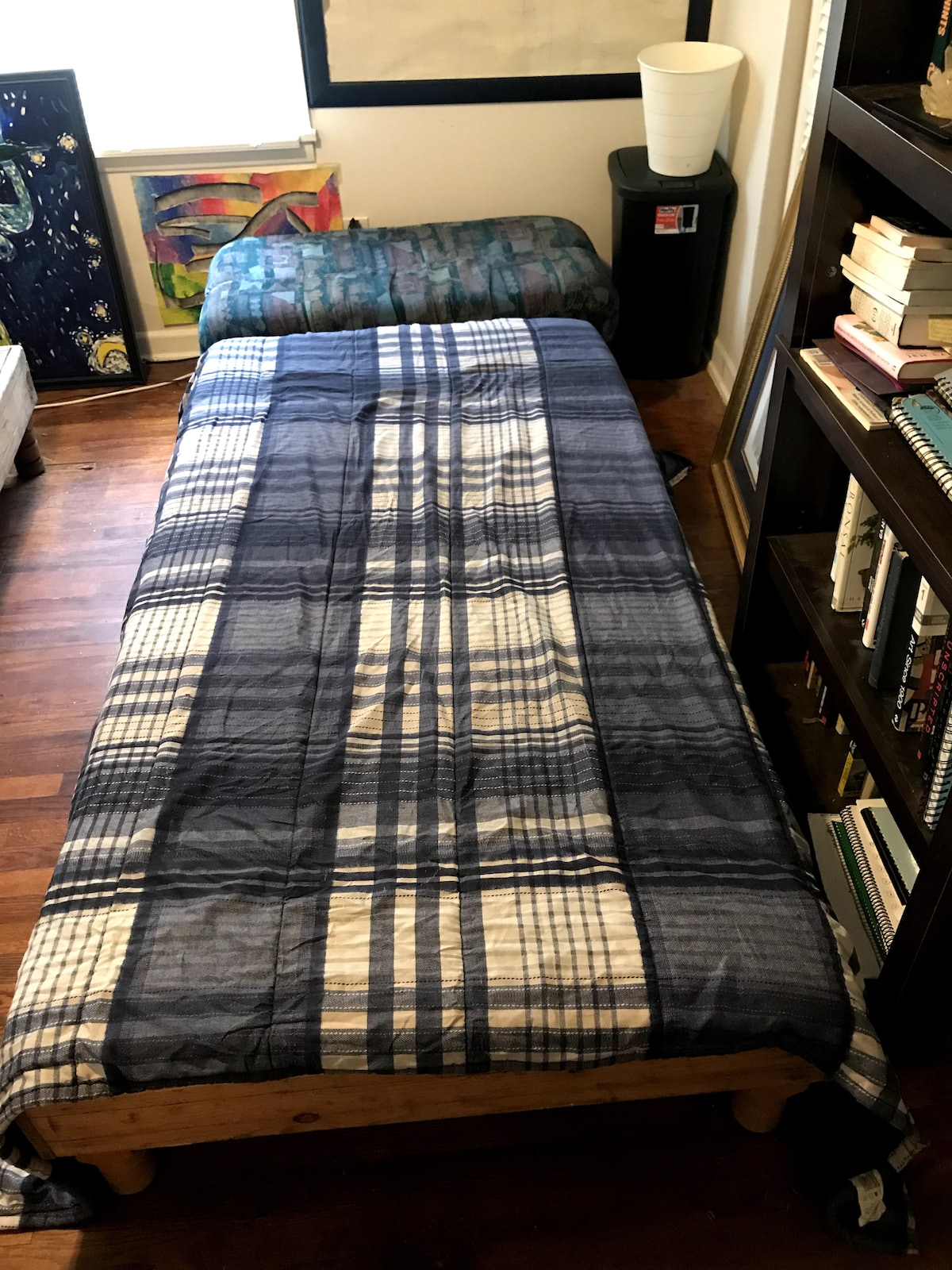 A cozy sleeping area is presented with a single bed covered in a blue plaid quilt. A small mattress is positioned nearby. Dark wooden flooring complements the workspace featuring a bookshelf filled with books and decorative items.