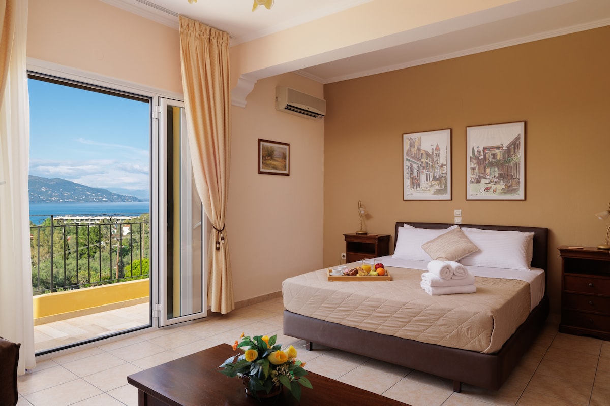 A spacious bedroom features a cozy double bed with fresh linens, situated near a set of glass doors opening to a private balcony that offers panoramic sea views. Light-colored walls and decor create a calming atmosphere, complemented by artwork displayed on the walls.
