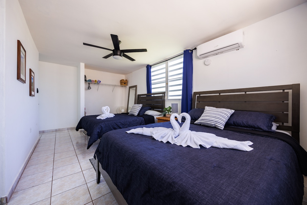 A well-lit bedroom features two spacious beds adorned with dark linens and decorative towel art shaped like swans. A ceiling fan is mounted above, and natural light enters through window blinds. Accessories are neatly arranged on a nearby shelf, complementing the simplicity of the space.