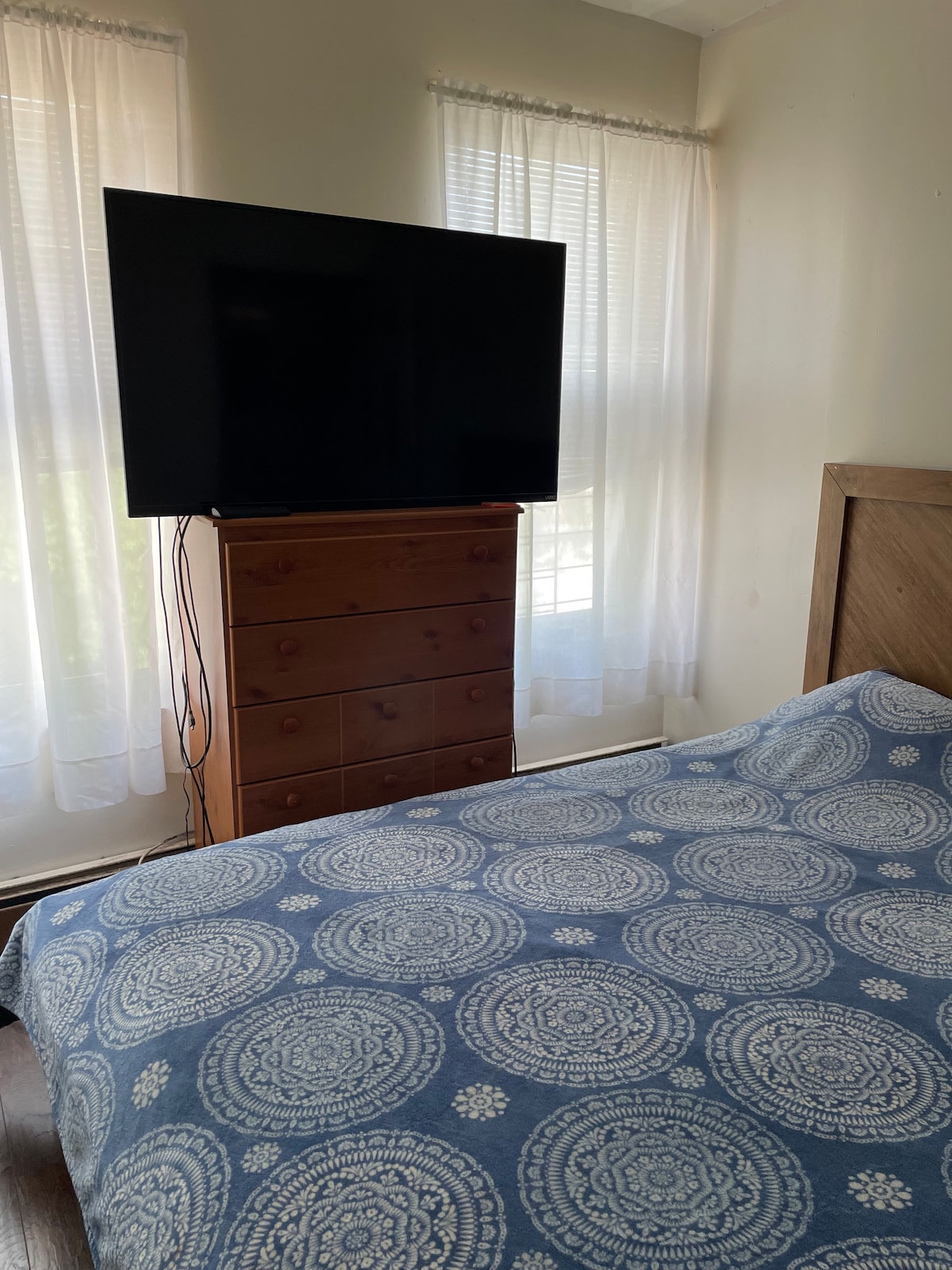 A bedroom features a large television mounted above a wooden dresser. Natural light filters through two sheer curtains, illuminating the blue patterned bedding on a spacious bed. The warm wood tones of the furniture create a calming ambiance.
