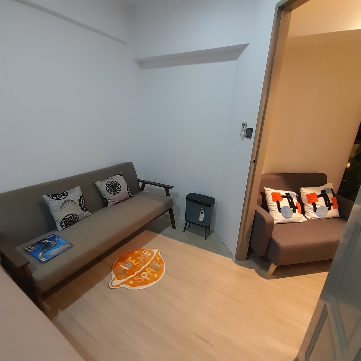 A welcoming seating area is visible, featuring a gray sofa embellished with decorative cushions. A small blue trash can is positioned nearby. A soft rug labeled 'Living Space' adds a touch of comfort, while a cozy armchair enhances the room's inviting atmosphere.