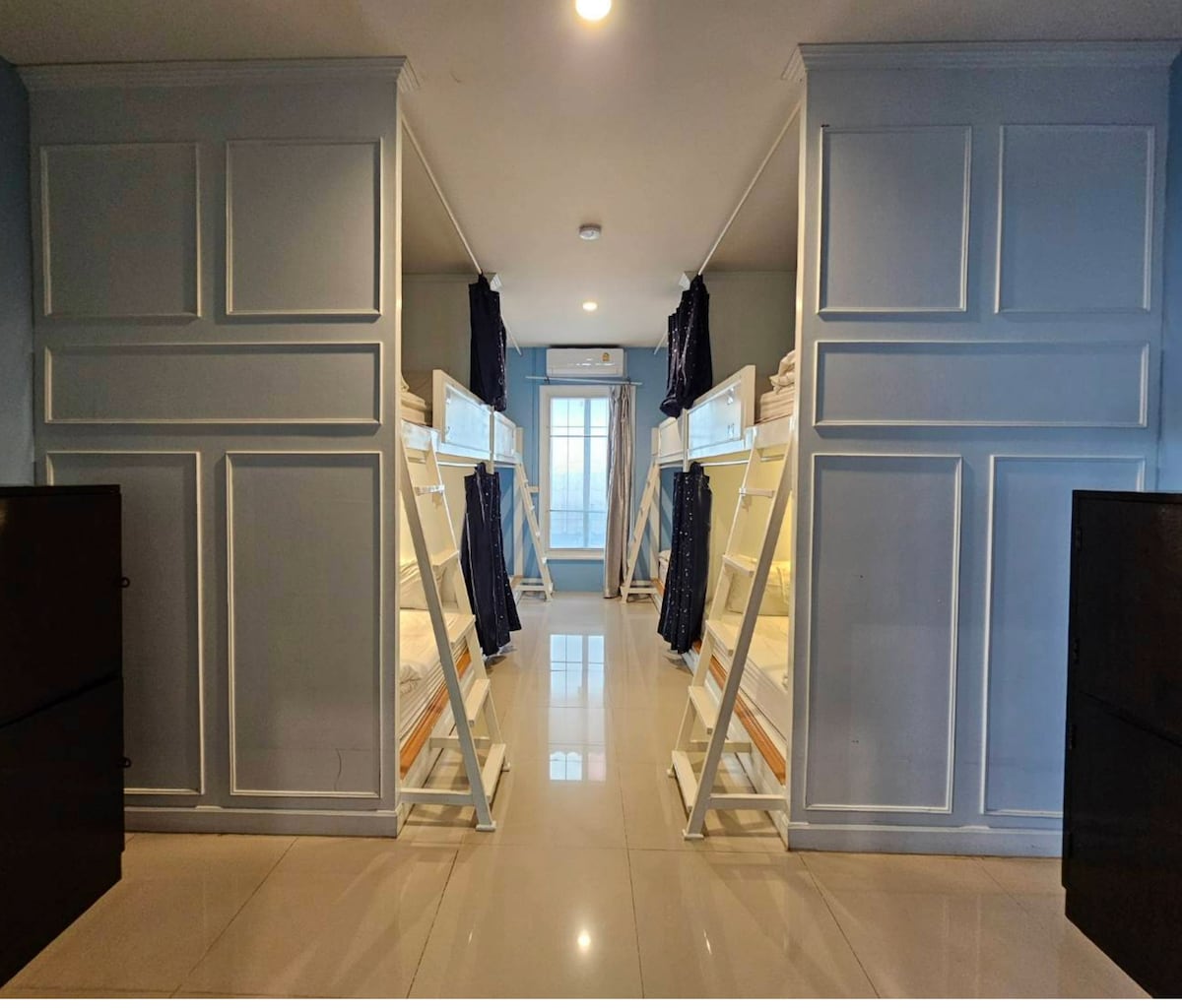 An open room features two sets of sturdy bunk beds, providing multiple sleeping spaces. The walls are painted in a soft blue, complemented by light-colored flooring. Natural light filters in through a window at the end of the corridor, flanked by dark curtains.