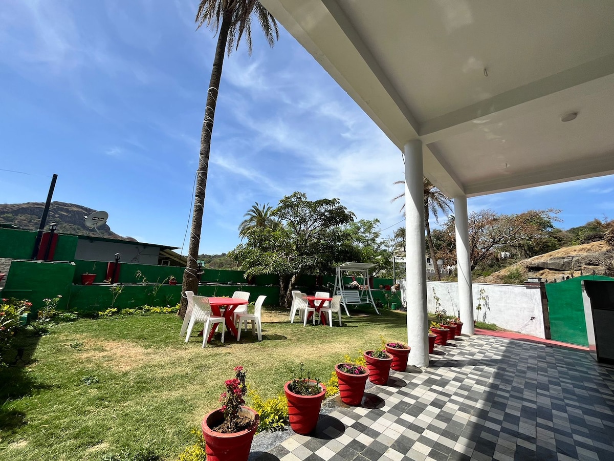 An outdoor area is featured with several white chairs and tables surrounded by green grass. Potted plants line the path, and a swing set is visible in the background. Palm trees provide shade, enhancing a relaxed environment for families.