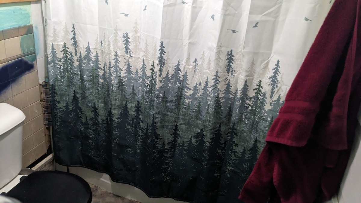 A bathroom is presented, featuring a forest-themed shower curtain with tall trees and flying birds. A toilet and a black chair are visible. Plush red towels add a touch of color, complemented by textured tile in natural hues on the wall.