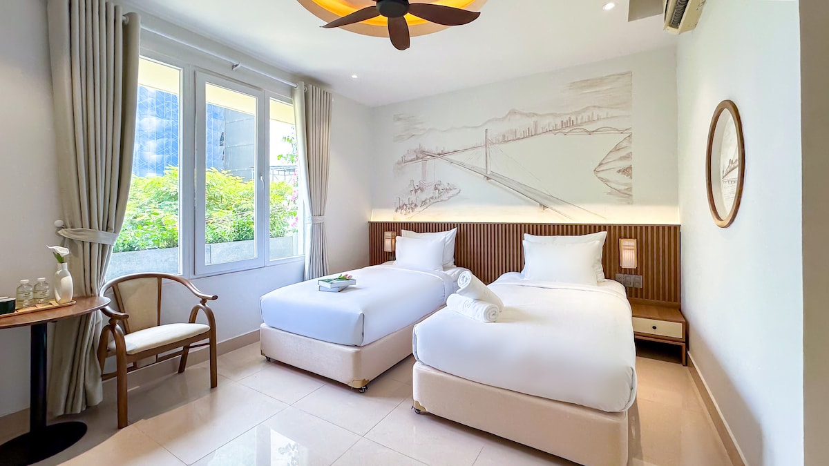 A modern guest room features two spacious beds with crisp white linens. Natural light filters through large windows, illuminating the space. A small wooden table is set beside a light-colored chair, and a wall art piece depicting a bridge adds a subtle touch to the decor.