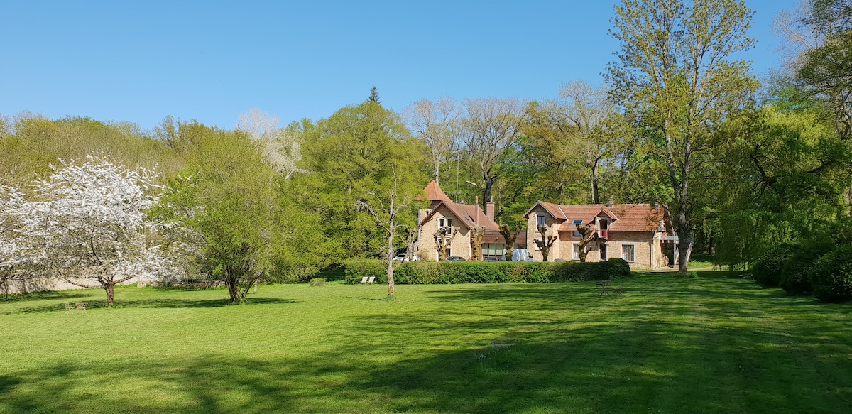 A vast green lawn is surrounded by trees and bushes, showcasing a natural and peaceful setting. The charming stone building with a red-tiled roof is visible in the background, blending harmoniously with the landscape. Flowering trees add a touch of seasonal beauty.