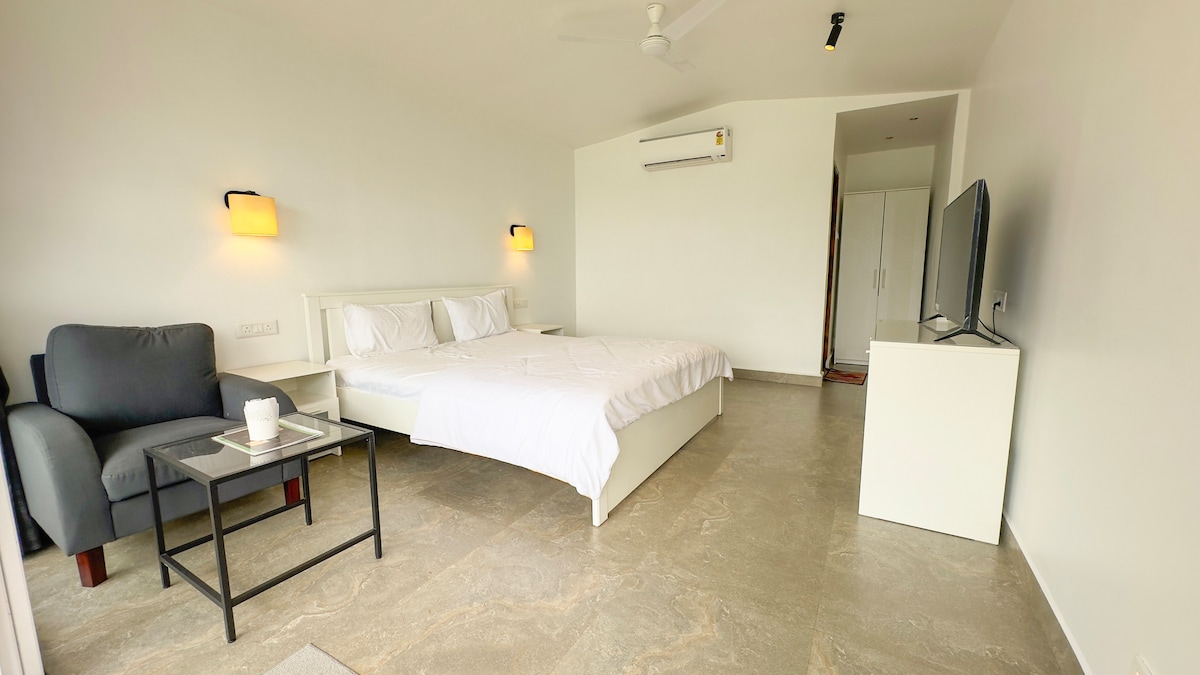 A bright and airy sleeping area features a spacious bed with pristine white linens. A comfortable armchair is positioned beside a small table. A flat-screen television is mounted on a white cabinet, and light fixtures on the walls provide a soft glow.