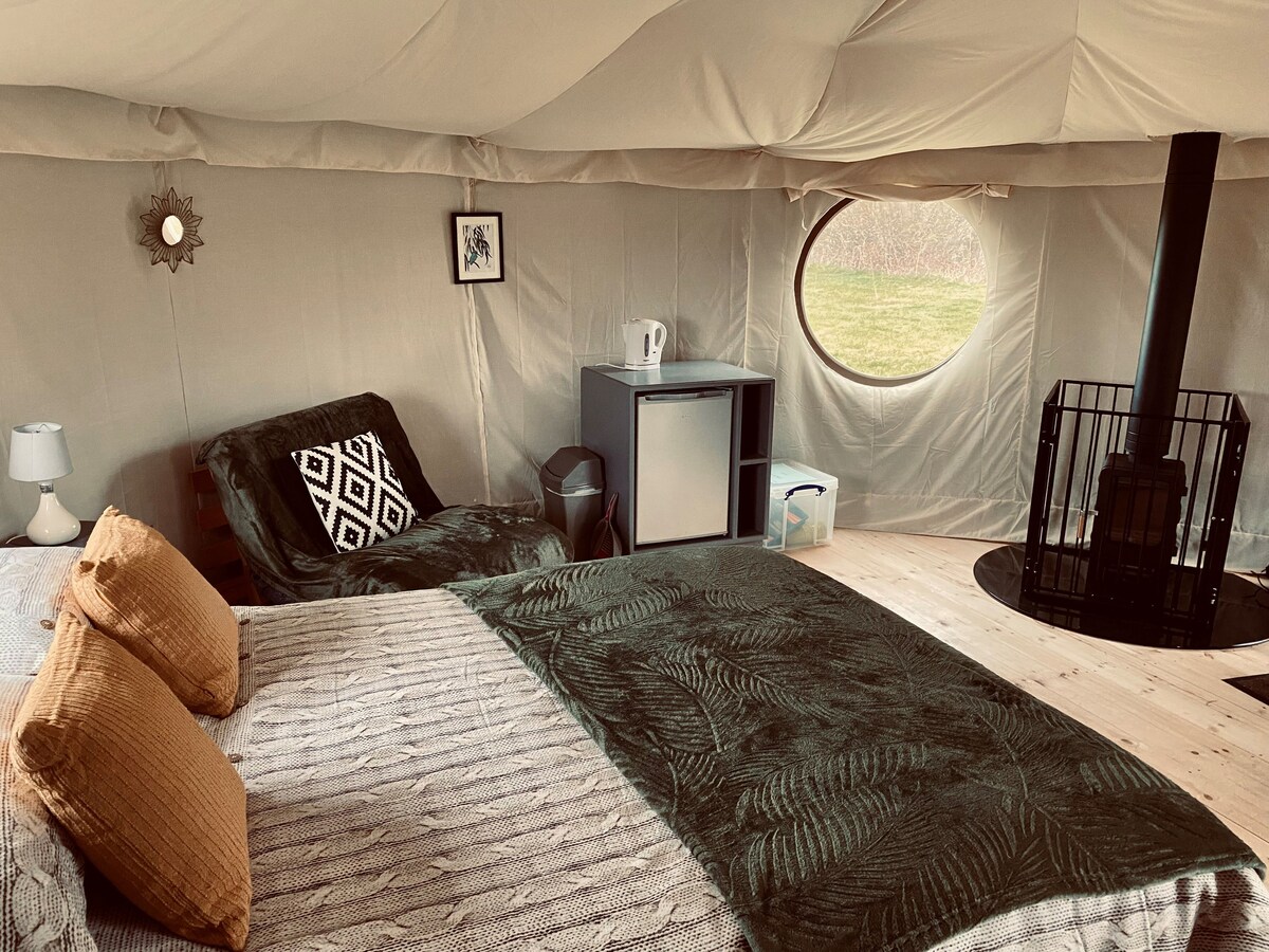 A spacious glamping tent interior features a double bed with decorative pillows and a cozy blanket. A comfortable chair is positioned next to a small fridge and a kettle. A log burner enhances the warmth, with an elegant round window providing natural light.