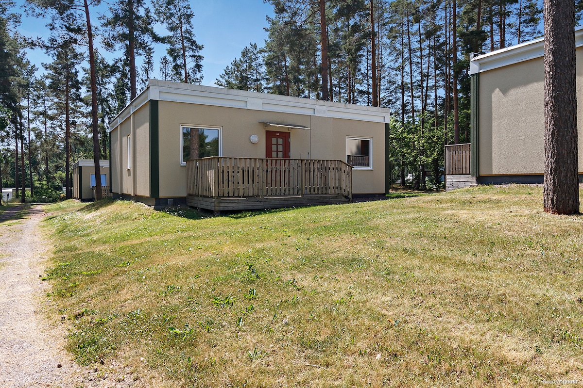 A spacious cabin is surrounded by trees, featuring a large wooden deck at the front entrance. The building has a simple facade with large windows, allowing natural light to enter. A manicured lawn leads up to the cabin, creating a welcoming outdoor space.
