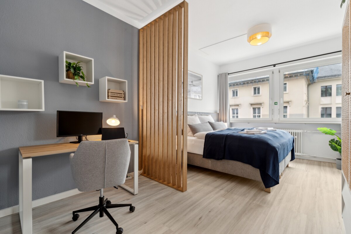 A modern bedroom features a comfortable bed with a blue blanket, separated from a workspace by a wooden screen. A desk with a chair and a monitor is positioned beside a window, allowing natural light to illuminate the room. Simple decor elements add to the inviting feel.