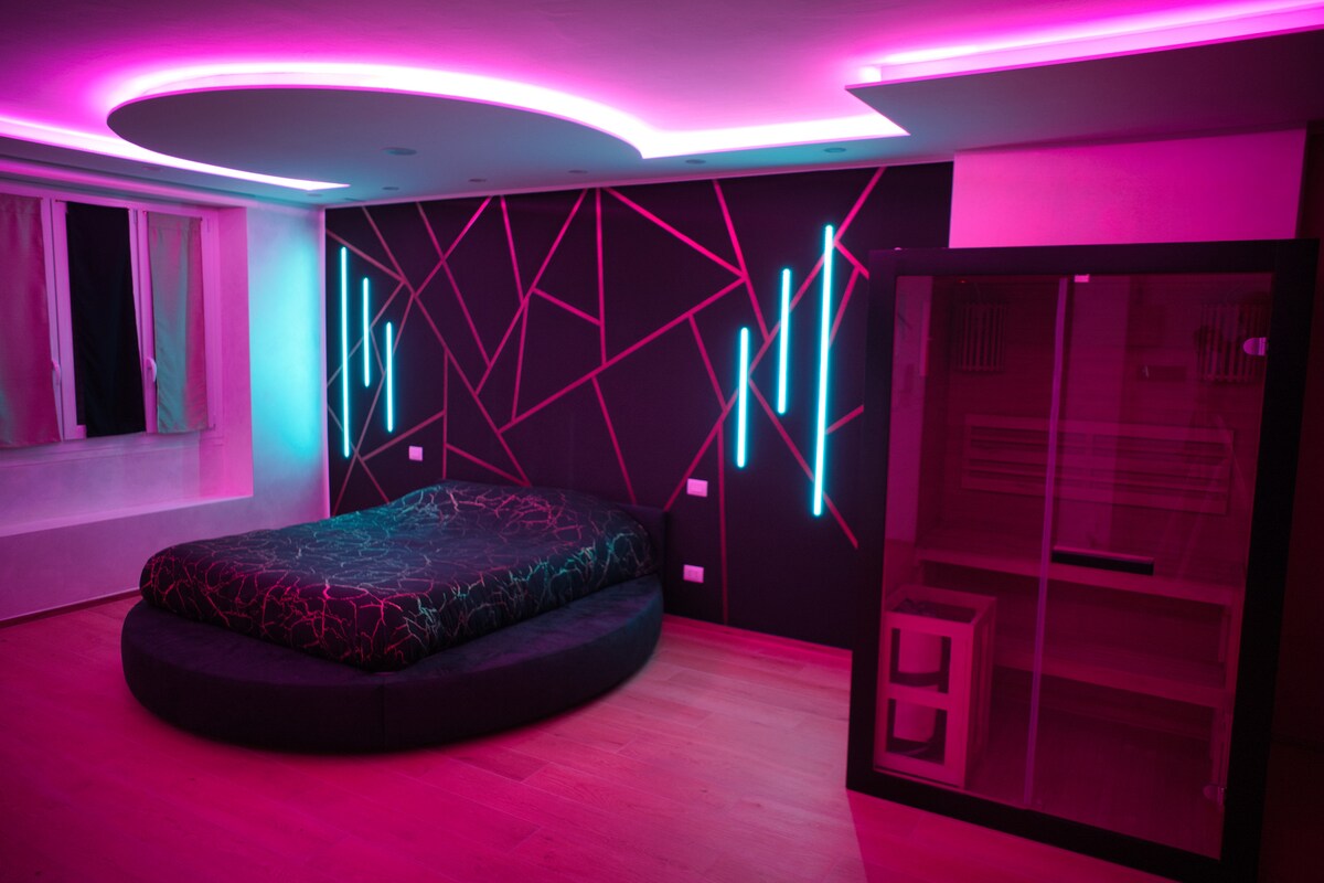 An elegant sleeping area features a spacious round bed with a dark patterned duvet. A dramatic backdrop showcases modern geometric wall designs illuminated in shades of pink and blue. A glass enclosure is visible, which likely houses a sauna, contributing to the room's luxurious atmosphere.