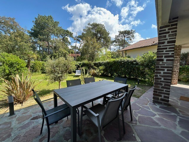 A spacious outdoor terrace is presented with a large dining table surrounded by six chairs. Lush greenery and well-maintained shrubs create a tranquil environment. A stone pathway leads to the house, while the sky reveals a mix of clouds and blue, enhancing the serene setting.