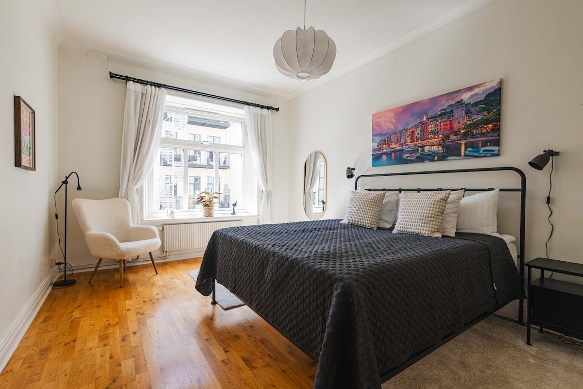 A spacious bedroom features a king-size bed with quality linens and decorative pillows. Natural light fills the room through a large window, which is framed by sheer white curtains. A cozy chair and side table are positioned near the window, paired with a mirror reflecting the room's design.