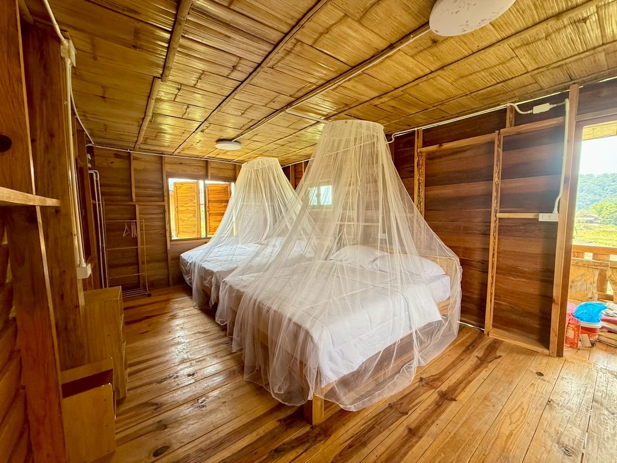 A spacious room features two beds draped in sheer mosquito nets. Natural wood elements are visible in the flooring and walls, complemented by ample natural light streaming through multiple windows. The serene atmosphere is enhanced by the rustic design and warm wood tones.