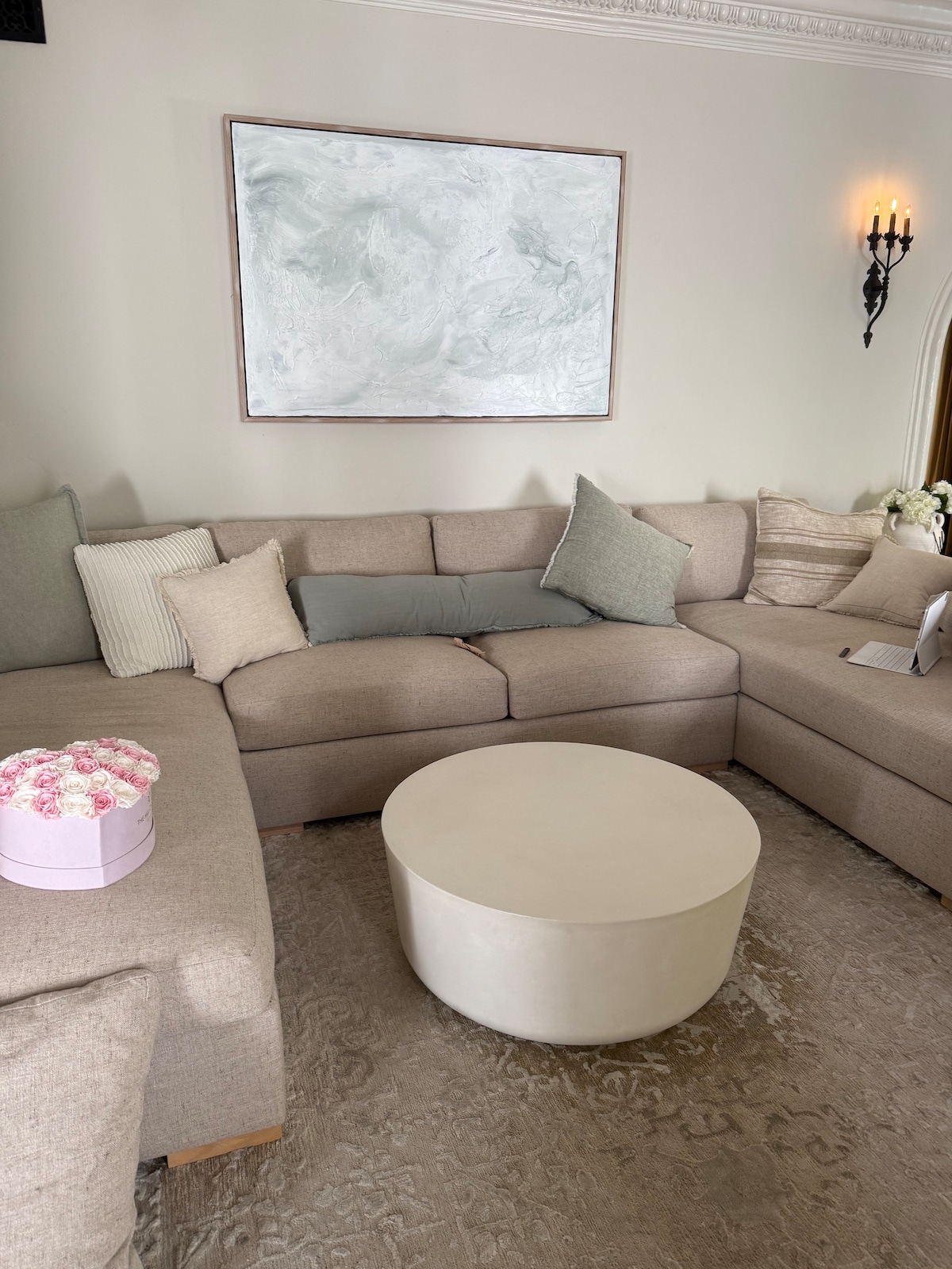 A comfortable seating area with a spacious sectional sofa in neutral tones is presented. A round, light-colored coffee table occupies the center space. The wall is adorned with a large abstract painting, and soft decorative pillows add subtle texture to the setting.