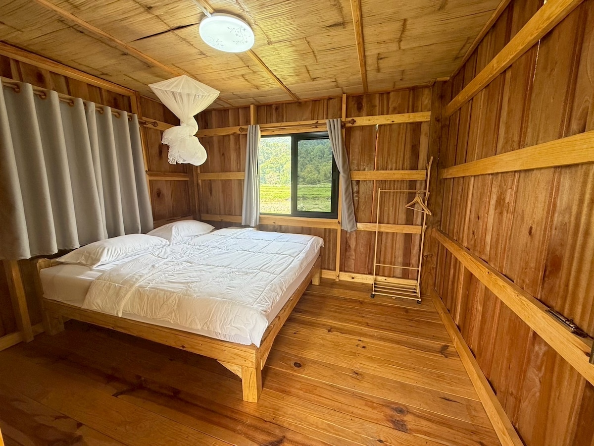 A rustic wooden interior features a spacious bed with crisp white bedding. Natural light enters through a large window, framed by light gray curtains. A minimalist clothes rack is positioned near the wall, complementing the warm tones of the wooden walls and flooring.