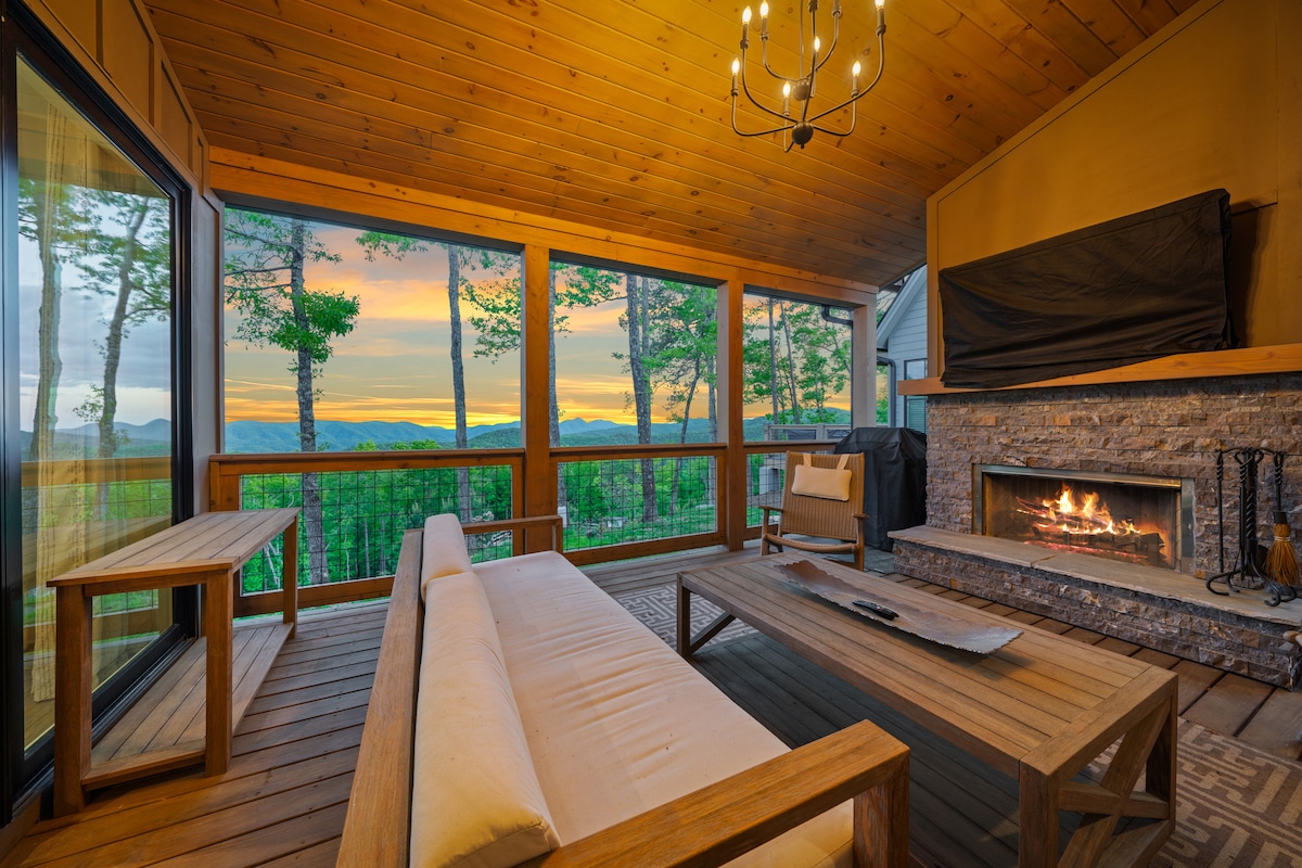 A cozy outdoor patio is shown with a wooden sofa and a rustic coffee table. A stone fireplace is visible, complementing the natural wood ceilings. Large glass windows frame the expansive mountain view and sunset, creating a serene space for relaxation.