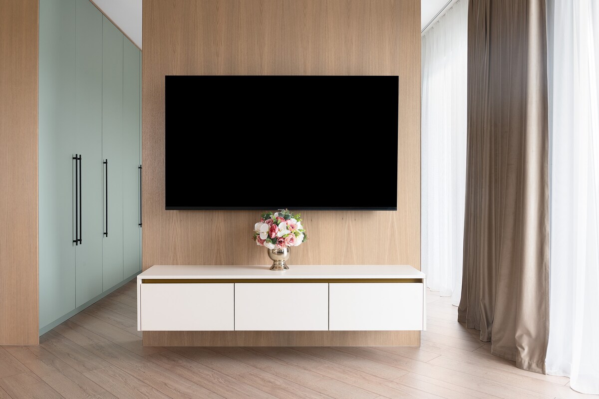A sleek white television unit, equipped with drawers, is positioned against a wooden wall. Above it, a flat-screen TV is mounted, providing a central focal point. A vase of flowers adds a touch of colour, while light enters through large curtains, enhancing the space's brightness.