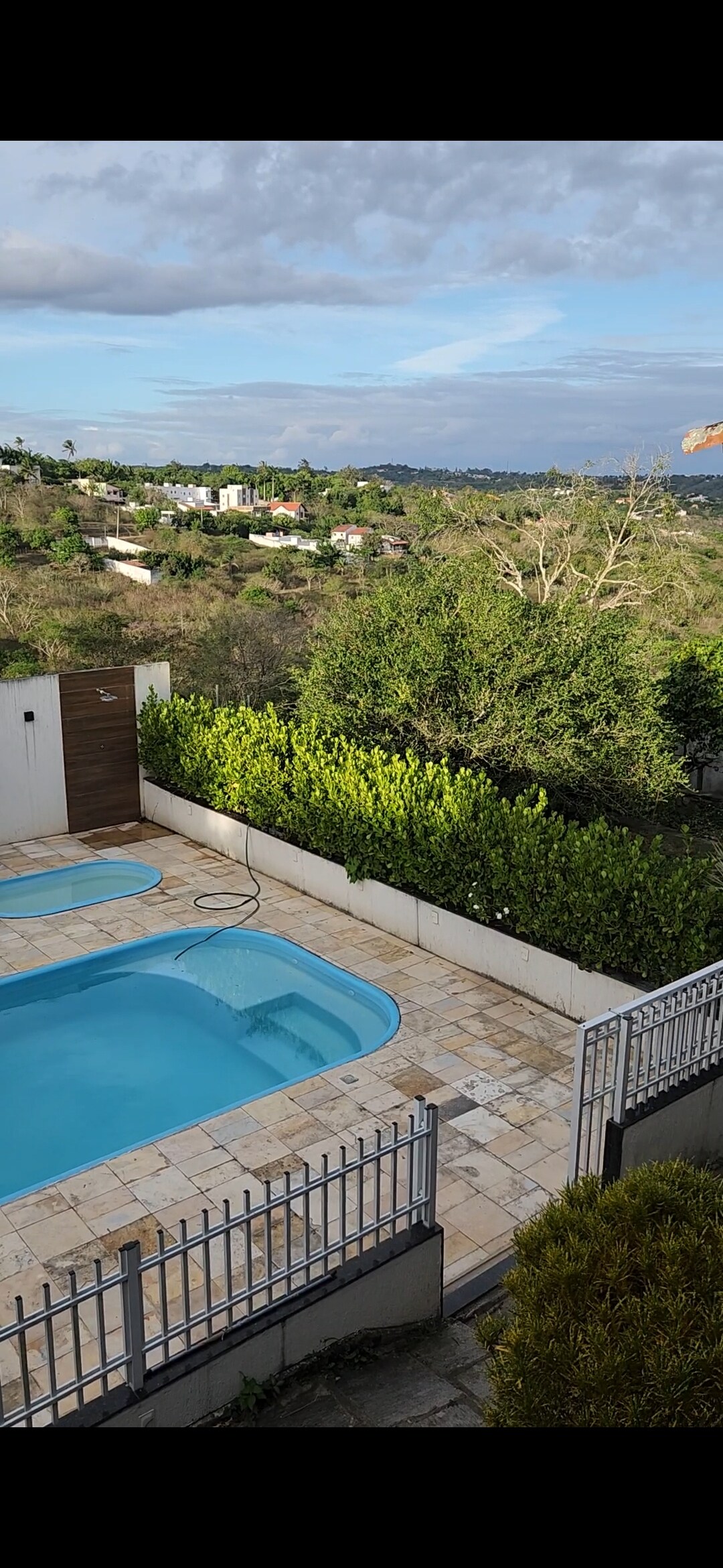 A pool area is featured, surrounded by greenery and a low fence. The pool, with a gentle curve, is set against a backdrop of rolling hills and houses in the distance. Clear skies are visible above, enhancing the serene atmosphere.