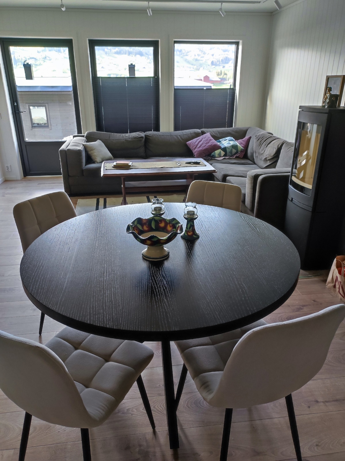 A round dining table with four upholstered chairs is positioned at the center. A small decorative bowl is placed on the table. In the background, a comfortable sofa is arranged with colorful cushions, and large windows allow natural light to fill the space.