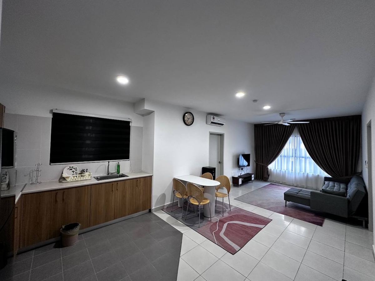 A spacious living area is featured, showcasing a kitchen with wooden cabinetry and a dining table surrounded by four chairs. A comfortable sofa is positioned in front of a television, while natural light enters through large windows adorned with dark curtains.
