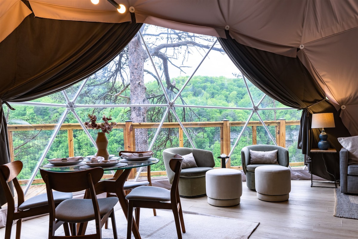 A dining area is visible, featuring a round table set for four, surrounded by wooden chairs. Two plush armchairs and matching ottomans offer comfortable seating. Large windows provide expansive views of greenery outside, enhancing the bright and airy ambiance.