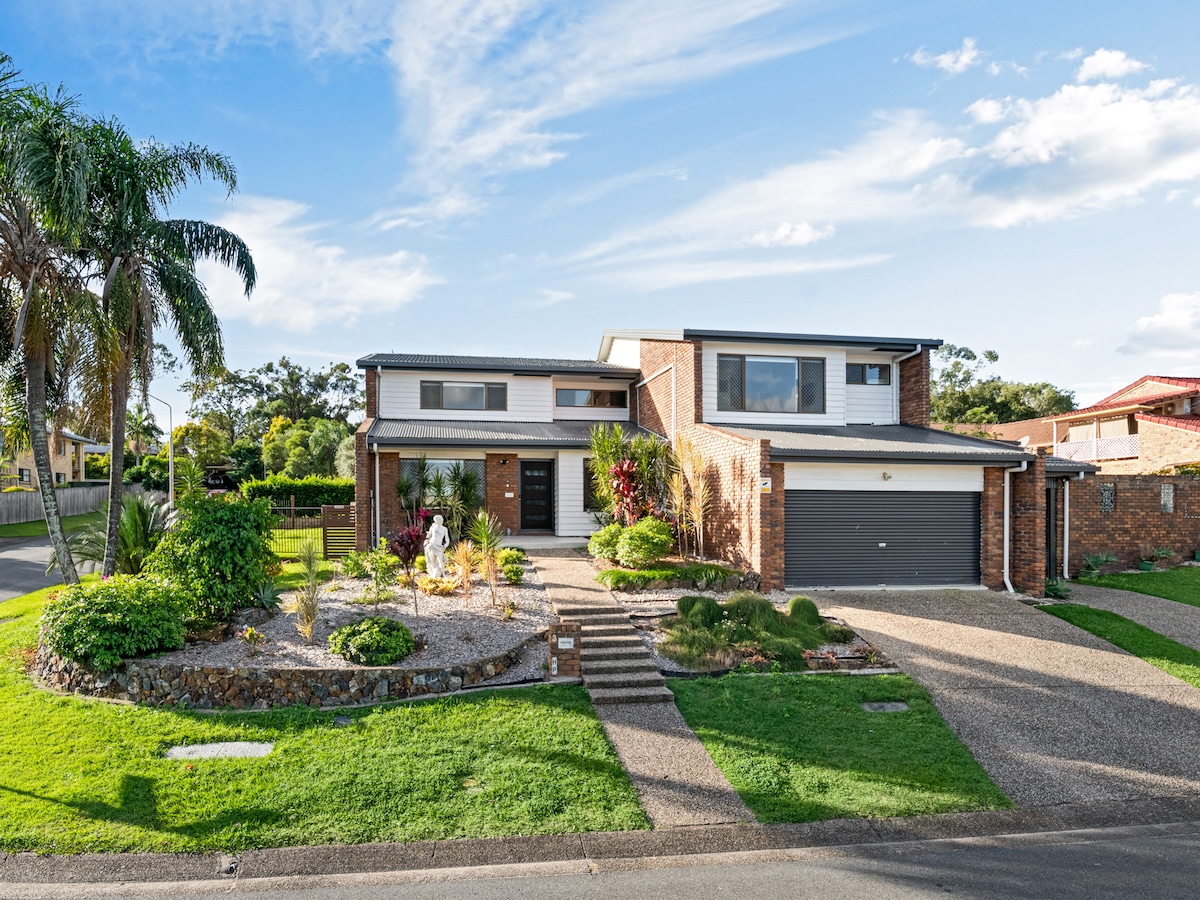 The exterior of a two-story residence is showcased, surrounded by manicured lawns and vibrant landscaping. Palm trees and shrubs frame the entrance, leading up to a pathway of steps. A double garage complements the brick facade, under a bright blue sky with scattered clouds.