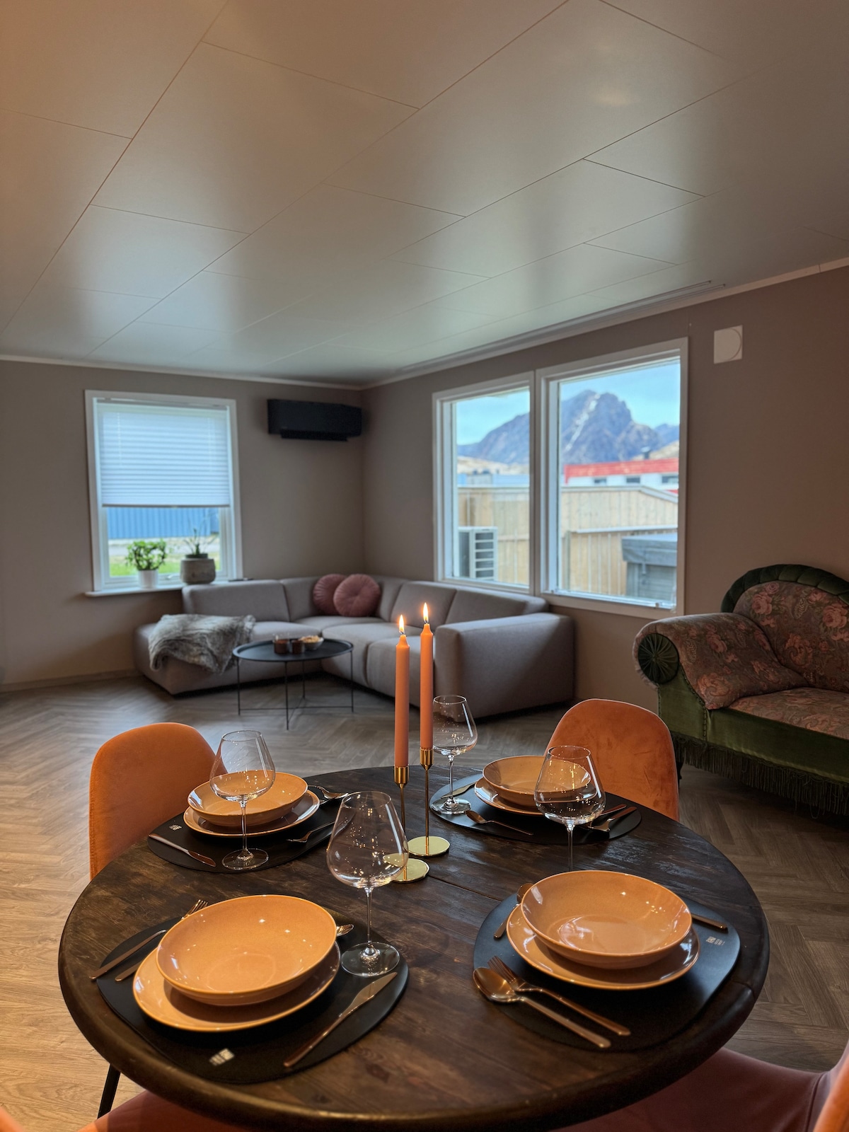 A dining area features a circular wooden table set with plates and wine glasses, complemented by two candles at the center. A comfortable sofa is positioned nearby, with large windows providing views of the surrounding landscape.