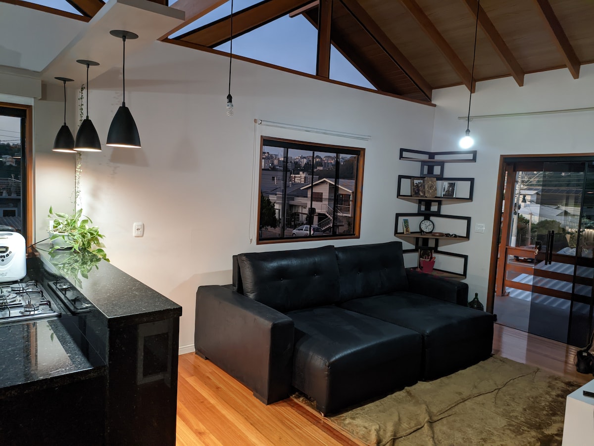 A modern living area features a sleek black sectional sofa complemented by warm wooden flooring. Large windows allow natural light to fill the space, while pendant lights hang elegantly above the adjacent kitchen area. A decorative shelving unit adds functionality and style.