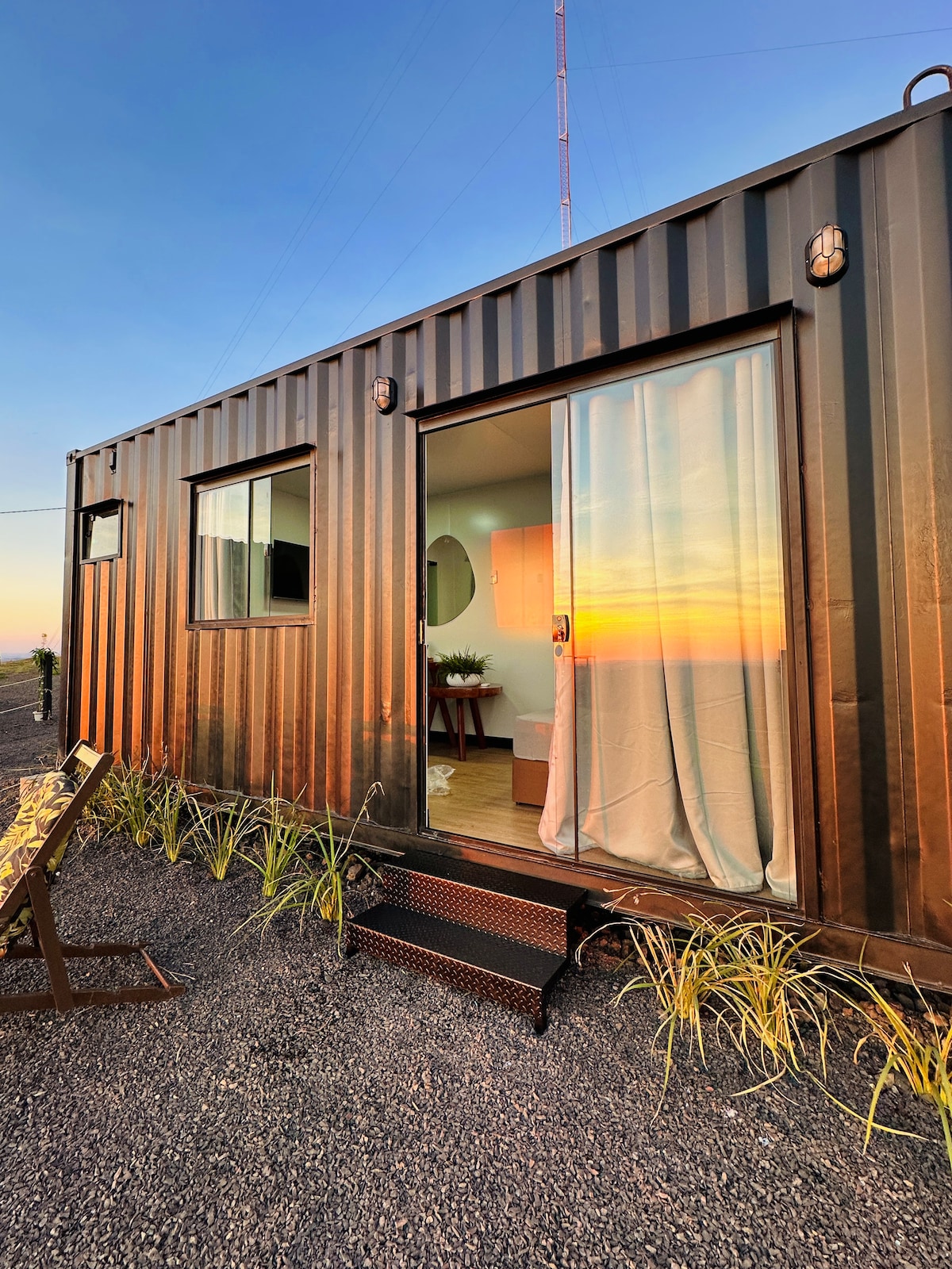 A sleek black container accommodation is presented, with large glass doors reflecting a vibrant sunset. Soft curtains frame the entrance, while a small staircase leads to the door. Surrounding greenery adds a touch of nature to the minimalist exterior.