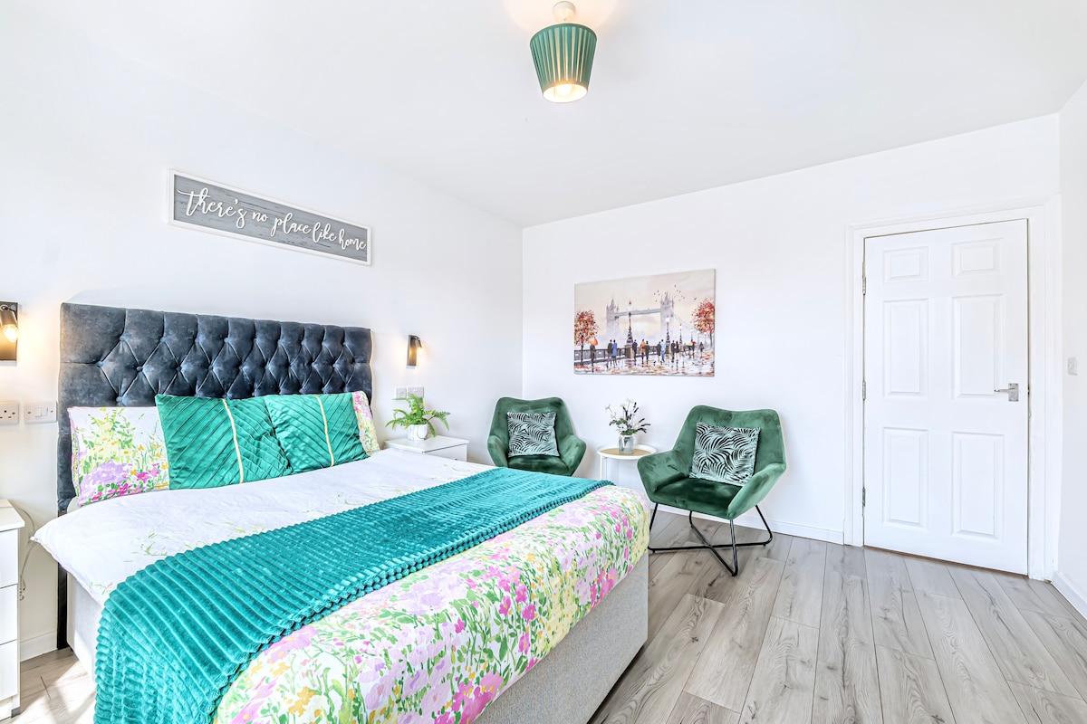 A modern bedroom features a spacious bed adorned with a floral print duvet and decorative pillows. Two green accent chairs are positioned beside a small plant, while a piece of wall art depicting a city scene is mounted above the bed. Natural light illuminates the room.