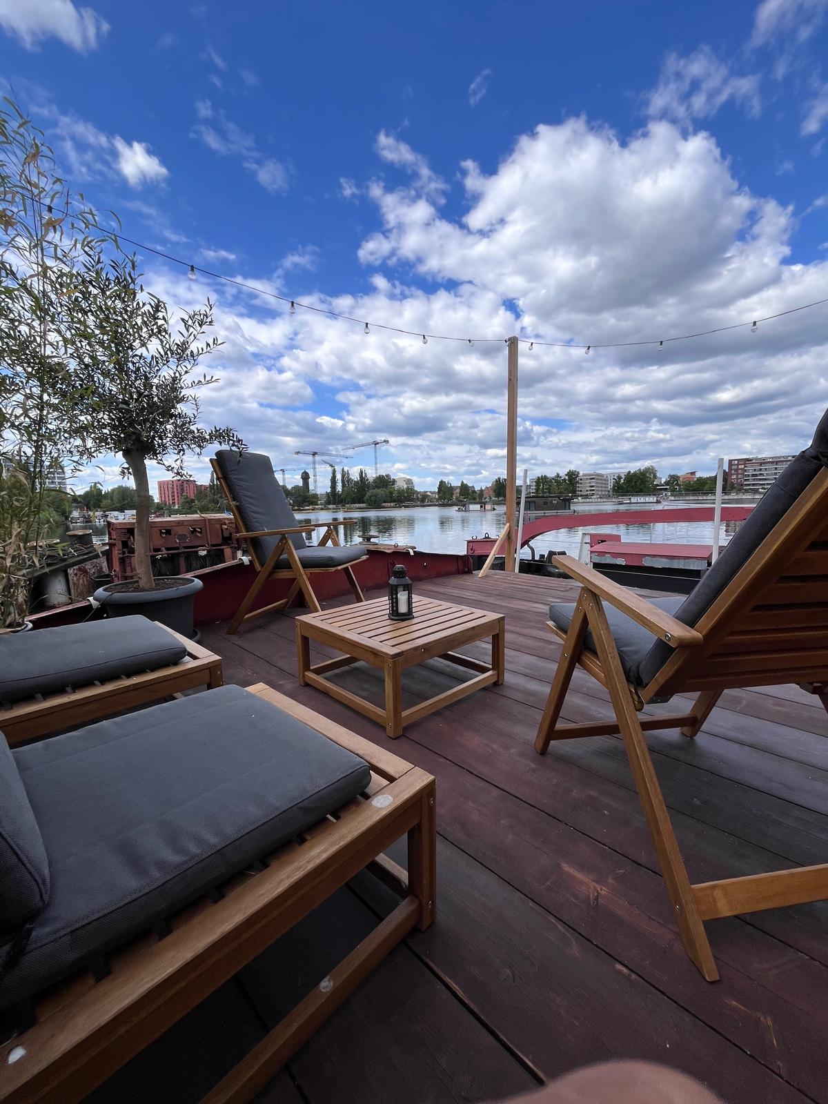 An outdoor sun deck features two lounge chairs with gray cushions positioned alongside a small wooden table. The area offers unobstructed views of the water and surrounding scenery, framed by a wide blue sky with scattered clouds.