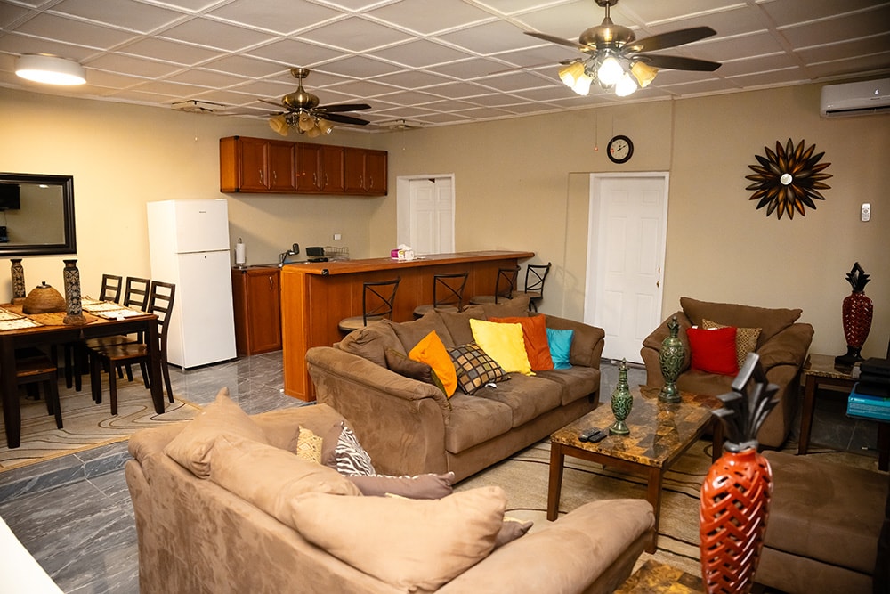 A spacious living area is furnished with brown sofas accented by colorful cushions. A wooden dining table with chairs is positioned near the kitchen bar. The kitchen features wooden cabinets and modern appliances, while overhead fans mounted on the ceiling provide comfort.