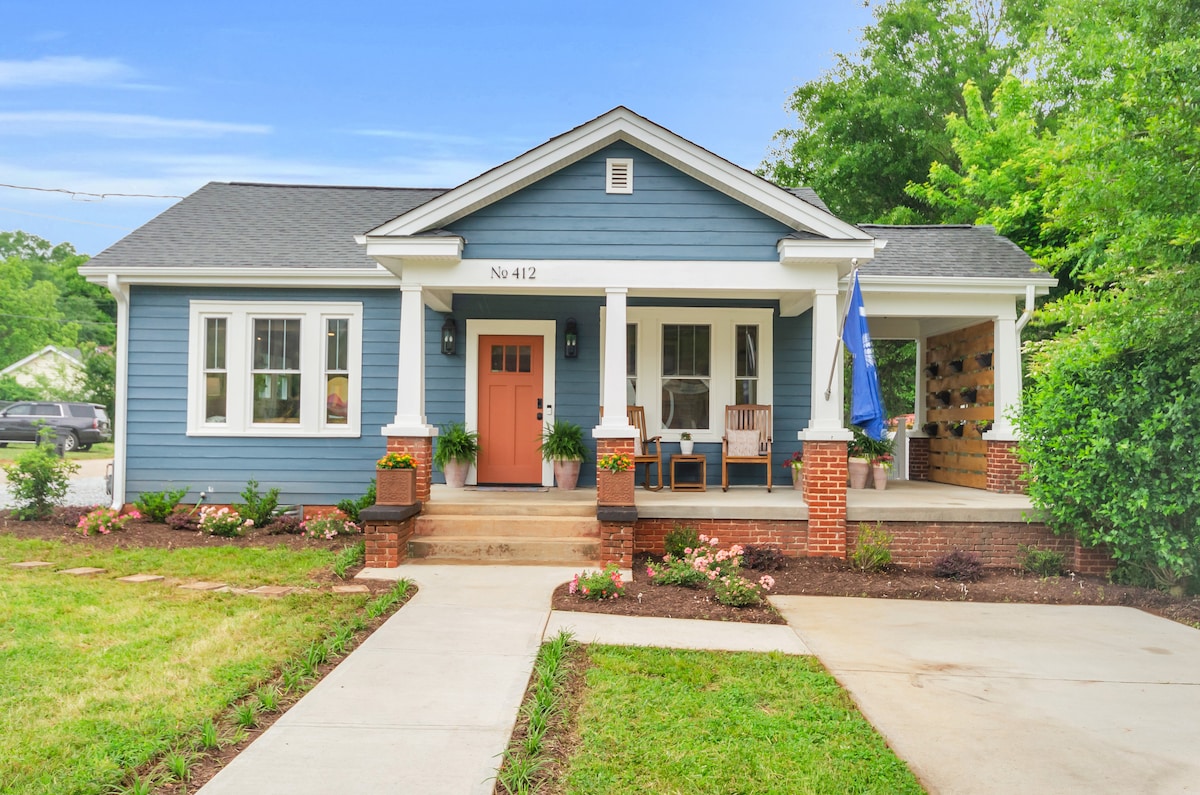 The exterior of The Botanical Bungalow is shown, featuring a charming blue facade with white trim. A welcoming front porch includes a couple of wooden rocking chairs and potted plants, while a pathway leads to the entrance. The surrounding yard is landscaped with greenery and seasonal flowers.