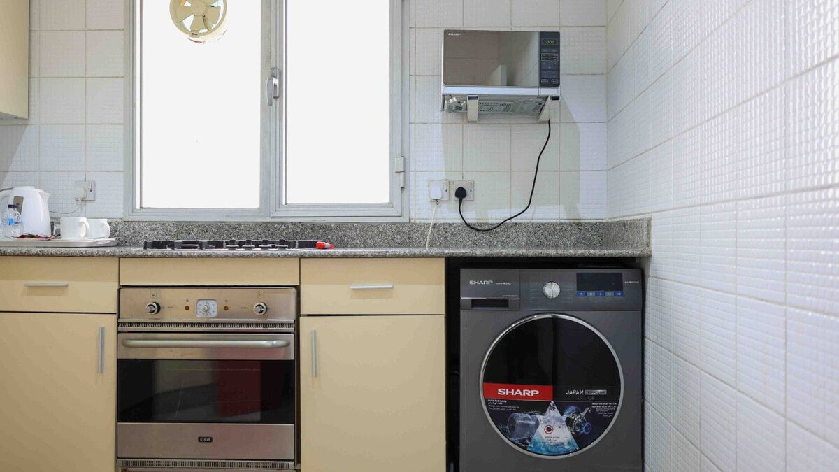 A modern kitchen is equipped with essential appliances, including a microwave and a washing machine. Light-colored cabinetry complements the granite countertops. A window allows natural light to fill the space, providing a view of the outside.