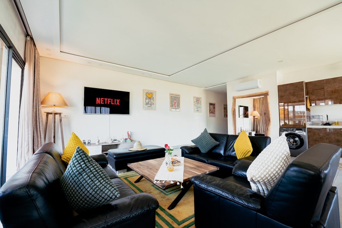 Détendez-vous sur un canapé en cuir confortable face à une télévision intelligente avec Netflix et des chaînes internationales. Le salon lumineux est décoré d'œuvres d'artistes bien connus, créant une atmosphère élégante, chaleureuse et inspirante. ✨