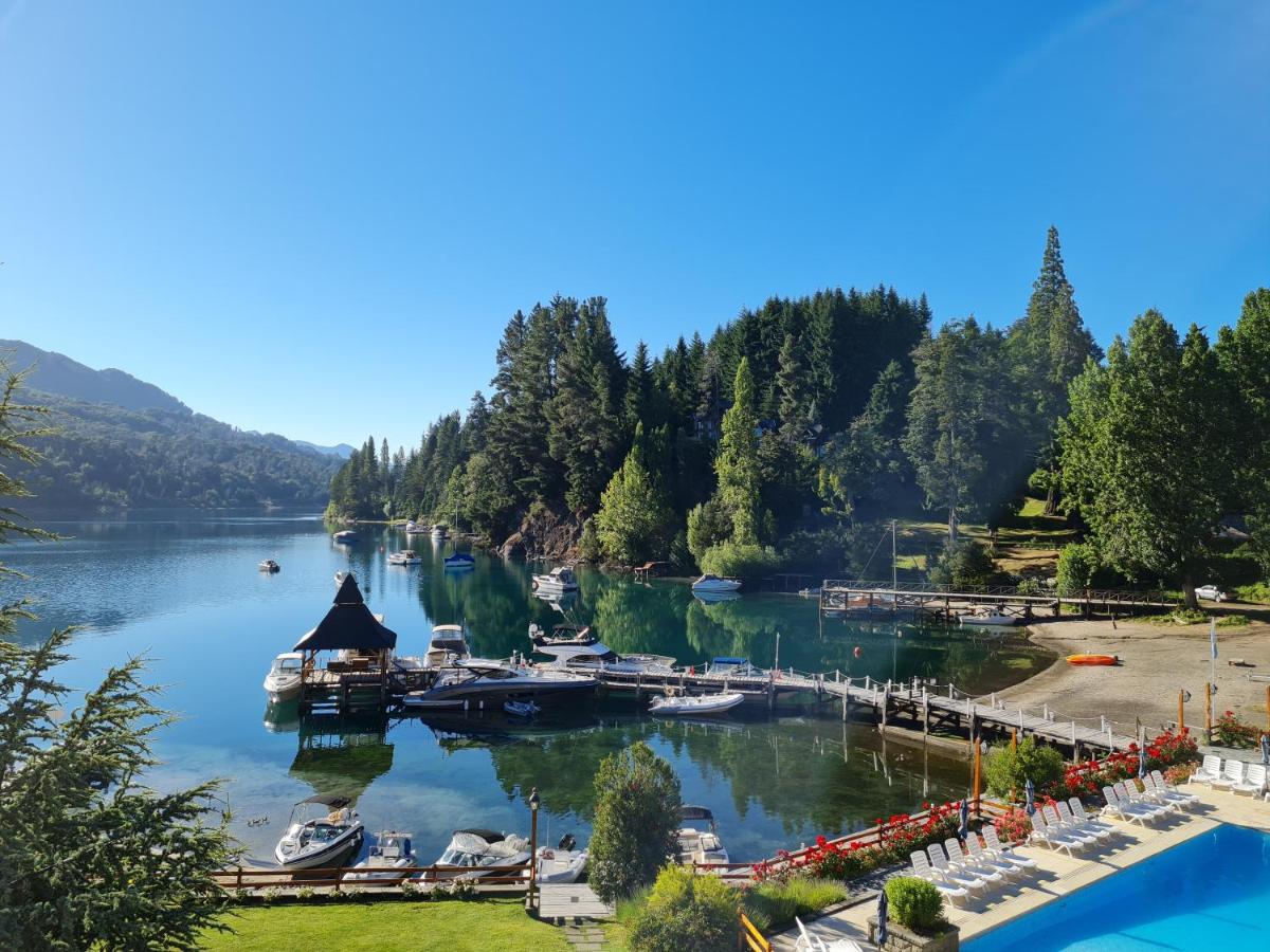 A serene bay on Lake Nahuel Huapi is visible, with calm waters reflecting the clear blue sky. Several boats are moored along the docks, surrounded by lush greenery and trees. The scene includes a small structure with a thatched roof, and sun loungers are placed near the water's edge.