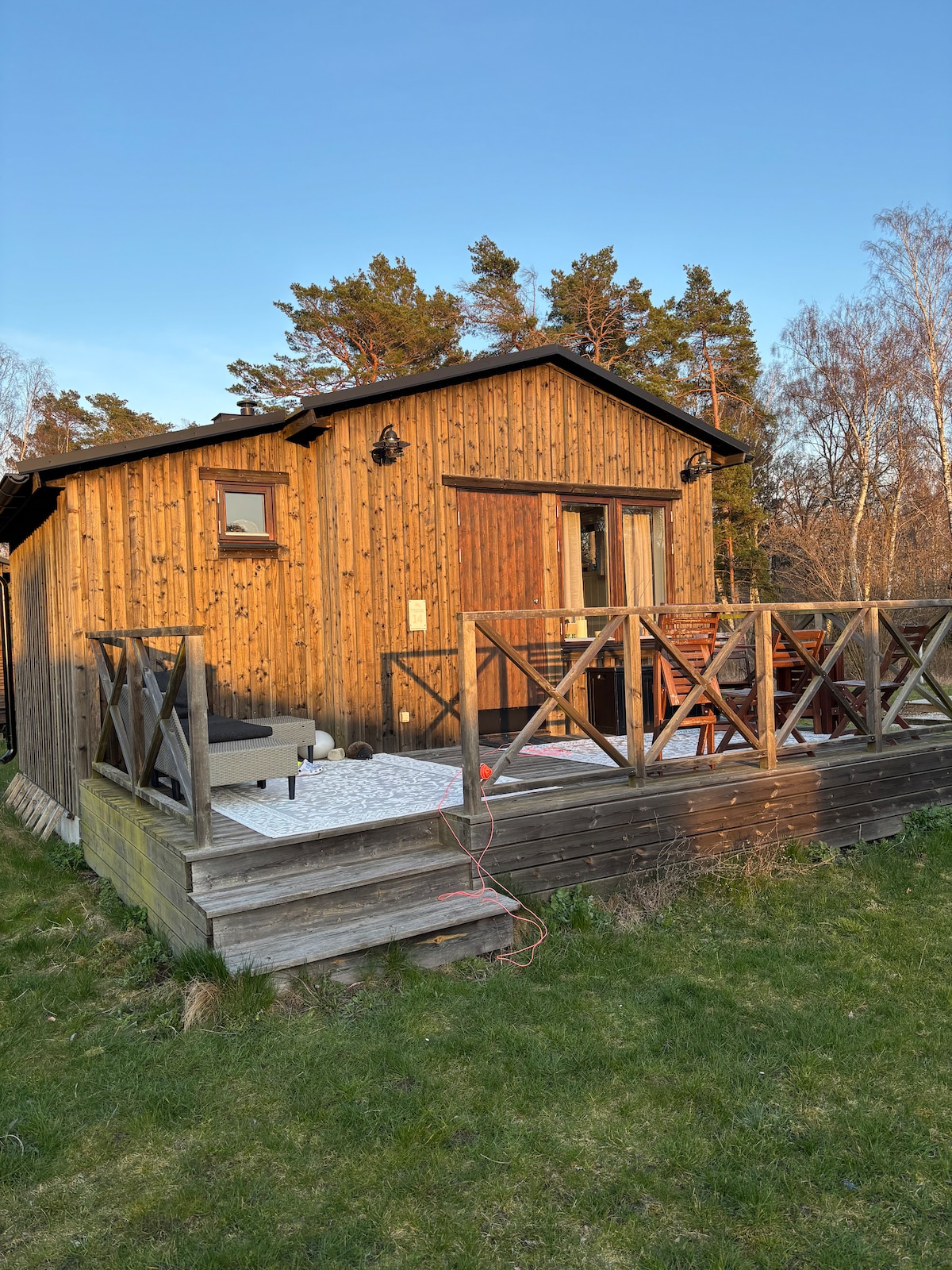 The exterior of a wooden cabin is showcased, featuring a front porch with a wooden railing. Outdoor seating is visible, along with grassy surroundings. Tall trees can be seen in the background under a clear sky, suggesting a serene and natural atmosphere.