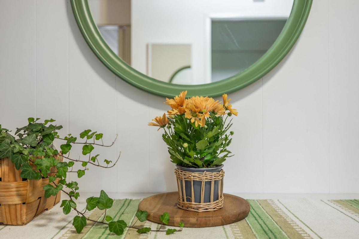 A green circular mirror is positioned above a wooden tray, highlighting a potted flowering plant with yellow blossoms. To the left, a woven basket contains greenery, and a patterned rug with green and white stripes completes the serene arrangement.