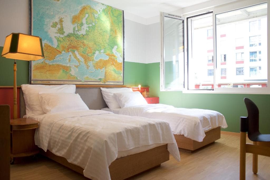 A bright sleeping area features two spacious beds with crisp white bedding situated against a green-accented wall. A large map of Europe is mounted above the beds. Natural light enters through a large window, supplemented by a simple lamp on a bedside table.