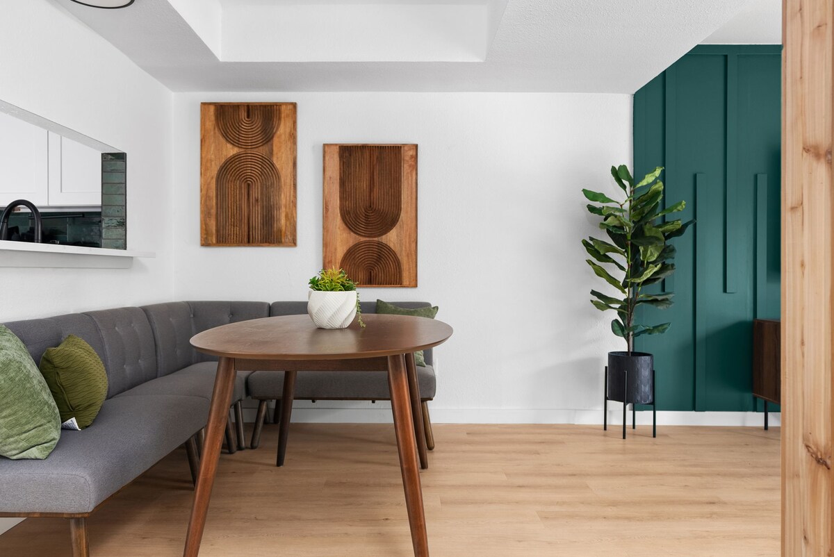 A modern dining area features a circular wooden table surrounded by a stylish gray sofa. Wooden wall art pieces provide texture against the white walls. A green leafy plant adds a natural touch, enhancing the overall calming ambiance of the space.
