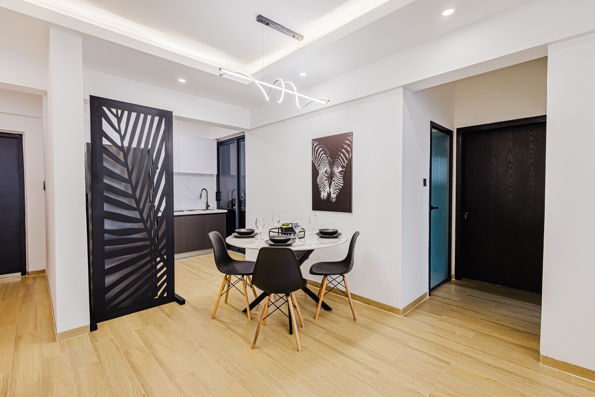 An open dining area features a round table set for four, accompanied by modern chairs. A decorative room divider adds an artistic touch, and the adjoining kitchen can be seen in the background. Neutral tones and contemporary design elements create a welcoming ambiance.